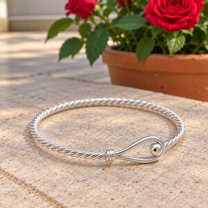 Puede incluir: Un brazalete de plata con una textura de cuerda trenzada en toda la banda. La pulsera incluye un mecanismo de cierre de tipo gancho con una bola de plata lisa y pulida que se asegura en un lazo en forma de gota en el extremo.