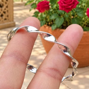 May include: A polished silver bangle bracelet featuring a continuous twisted, spiral design. The metal surface is smooth and reflective, catching the light along the curves of the circular band. The bracelet has a uniform thickness throughout its structure.