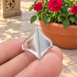 May include: A silver-toned ring featuring a prominent geometric design with a pointed, diamond-like shape. The surface of the ring displays a textured, matte finish across its triangular panels, creating a distinct contrast with the smooth, polished edges.