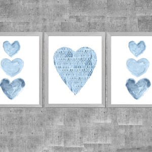 K&ouml;nnte beinhalten: Drei gerahmte Wandbilder mit blauen Herzmotiven. Die beiden &auml;u&szlig;eren Rahmen zeigen drei &uuml;bereinander angeordnete Herzen im Aquarellstil, w&auml;hrend der mittlere Rahmen ein gro&szlig;es Herz mit einem blauen schuppenartigen Muster zeigt.