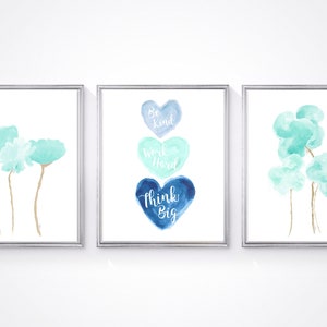 May include: Three watercolor prints in silver frames. The left print features two light blue flowers with gold stems. The center print features three watercolor hearts in shades of blue with the text "Be Kind, Work Hard, Think Big". The right print features three light blue watercolor clouds with gold stems.