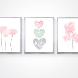 May include: Three watercolor prints in silver frames. The center print features three watercolor hearts with the words "Be Kind", "Work Hard", and "Think Big". The prints on either side feature pink watercolor flowers.