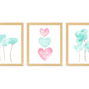 May include: Set of three framed watercolor prints with a light blue and pink color scheme. The first print features a cluster of light blue watercolor flowers. The second print features three stacked watercolor hearts with the words "Be Kind", "Work Hard", and "Think Big" written in white. The third print features a cluster of light blue watercolor clouds.
