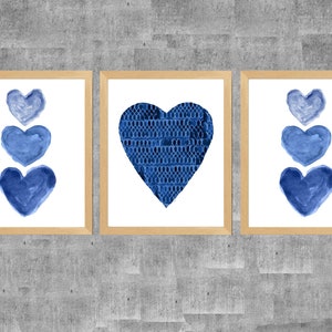 K&ouml;nnte beinhalten: Drei gerahmte Wandbilder mit hellen Holzrahmen. Die beiden &auml;u&szlig;eren Bilder zeigen jeweils eine vertikale Anordnung von drei blauen Aquarellherzen. Das mittlere Bild zeigt ein gro&szlig;es Herz mit einem dunkelblauen Spitzenmuster.