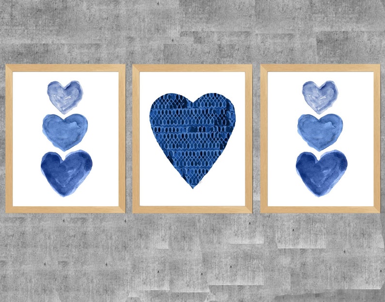 K&ouml;nnte beinhalten: Drei gerahmte Wandbilder mit hellen Holzrahmen. Die beiden &auml;u&szlig;eren Bilder zeigen jeweils eine vertikale Anordnung von drei blauen Aquarellherzen. Das mittlere Bild zeigt ein gro&szlig;es Herz mit einem dunkelblauen Spitzenmuster.