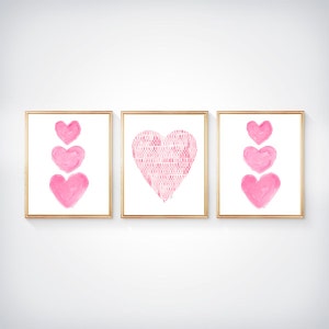 K&ouml;nnte beinhalten: Ein Set aus drei gerahmten Wandbildern. Die linken und rechten Bilder zeigen jeweils eine vertikale Anordnung von drei rosa Aquarellherzen. Das mittlere Bild zeigt ein gro&szlig;es Herz mit einem sich wiederholenden rosa-wei&szlig;en Muster.