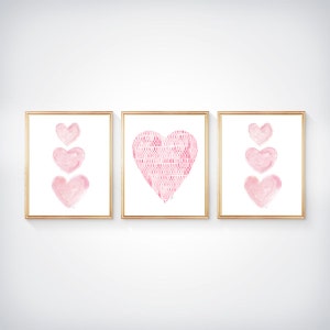 K&ouml;nnte beinhalten: Ein Set aus drei gerahmten Wandbildern mit rosa Herzillustrationen. Die beiden &auml;u&szlig;eren Rahmen zeigen drei &uuml;bereinander angeordnete Herzen im Aquarellstil, w&auml;hrend das mittlere Bild ein gro&szlig;es Herz mit texturiertem Muster zeigt.