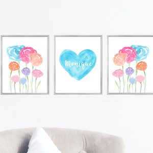 Könnte beinhalten: Set aus drei gerahmten Aquarelldrucken. Zwei Drucke zeigen bunte Blumen in Rosa, Orange, Blau und Lila. Der mittlere Druck hat ein blaues Herz mit dem Namen "Monique" in weißer Schrift. Die Drucke sind in silbernen Rahmen.