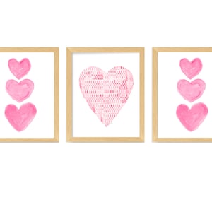 Könnte beinhalten: Drei gerahmte Drucke mit rosa Aquarellherzen. Der mittlere Druck zeigt ein großes Herz mit einem Muster. Die beiden anderen Drucke zeigen jeweils drei Herzen, die vertikal übereinander gestapelt sind.