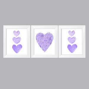 以下が含まれることがあります： 紫色の水彩画のハートが描かれた額装されたプリントが3枚。中央のプリントには、模様のある大きなハートが描かれている。他の2枚のプリントには、それぞれ3つのハートが縦に積み重ねられており、一番下のハートが最も大きい。