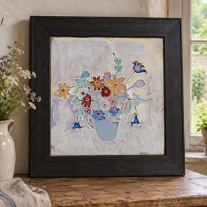 Op de afbeelding: Een vierkant ingelijst kunstwerk aan de muur met een kleurrijk bloemstuk in een lichtblauwe vaas. De bloemen zijn afgebeeld in tinten rood, oranje, geel en blauw tegen een gestructureerde, bleke, gemarmerde achtergrond. De lijst is van donker hout.