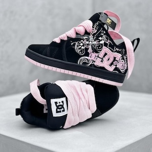 Puede incluir: Un par de zapatillas DC negras y rosas con cordones rosas gruesos, parte superior de ante negro y charol con patrones gráficos blancos, un logotipo DC rosa en el lateral y un dije de cruz plateada unido al ojal con una etiqueta negra.