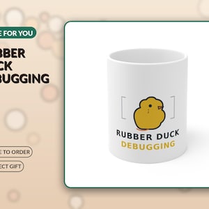 May include: A white ceramic mug featuring a graphic of a yellow rubber duck centred between two brackets. Below the duck, the text reads RUBBER DUCK DEBUGGING in black and yellow lettering. The mug has a smooth finish and a cylindrical shape.