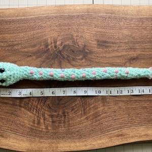 May include: A light mint green crocheted snake toy featuring black plastic eyes and small pink and purple beads embedded along its body. A white measuring tape placed underneath shows the snake has a total length of approximately 16 inches.