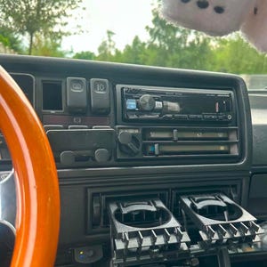 Könnte beinhalten: Ein Auto-Armaturenbrett mit einem schwarzen Alpine-Radio, einem hölzernen Lenkradkranz auf der linken Seite und zwei ausgefahrenen schwarzen Kunststoff-Getränkehaltern. Ein Paar flauschige weiße Würfel hängt über der Konsole.