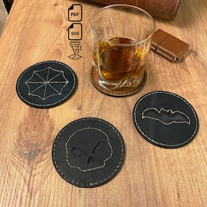May include: Three circular black leather coasters featuring laser-cut designs of a skull, a bat, and an umbrella-like shape. Each coaster has light-colored stitching around the perimeter. A clear glass with amber liquid sits on one coaster.