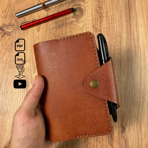 May include: A tan leather notebook cover with visible stitching, a brass-colored snap closure, and a black pen inserted into the side. Icons for PDF, SVG, a laser cutter, and a play button are visible, along with a silver and a red pen.