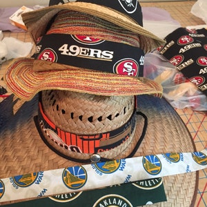May include: A stack of straw hats with different sports team logos. The top hat has a Raiders logo, the middle hat has a 49ers logo, and the bottom hat has a Warriors logo.