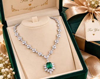 May include: A silver-toned necklace featuring a central emerald-cut green gemstone surrounded by marquise and round clear stones, with a leaf-patterned chain. The necklace is displayed in a dark green velvet box with the text A Special Gift For You.
