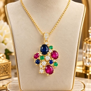 Puede incluir: Un collar de oro con un gran colgante que presenta un grupo de piedras preciosas ovaladas y redondas en azul oscuro, rojo vibrante y verde brillante, intercaladas con pequeñas piedras blancas claras. El colgante cuelga de una fina cadena.