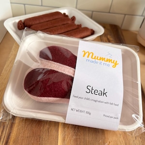 Puede incluir: Juego de comida de fieltro con un paquete de dos filetes de fieltro y una bandeja de salchichas. El paquete de filetes tiene una etiqueta que dice "Mummy made it me" y "Steak". El peso neto es de 100g.