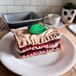 May include: A felt lasagna slice on a white plate. The layers include tan, red, and brown felt, with a green leaf on top. The lasagna has a ruffled edge. A baking pan, a cup and saucer, and a decorative plant are in the background.