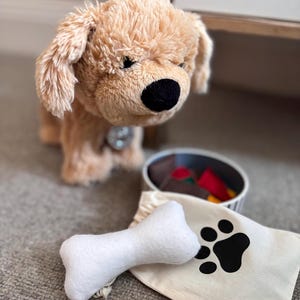 Puede incluir: Un juguete de peluche con forma de perro de color canela con una nariz negra y orejas caídas. Un juguete en forma de hueso blanco y una bolsa de lona con una huella de pata negra están frente a un cuenco con comida de juguete. Un juego de regalo con temática de mascotas.