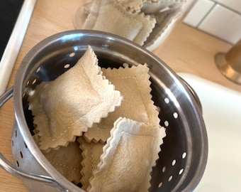 Può includere: Un colino in acciaio inox pieno di ravioli di feltro beige. I ravioli sono impilati uno sopra l'altro.