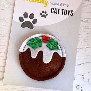 Può includere: Un giocattolo per gatti in feltro a forma di budino di Natale. Il budino marrone ha glassa bianca, foglie di agrifoglio verdi e bacche rosse. La carta dice "Mummy made it me CAT TOYS" con impronte di zampe.