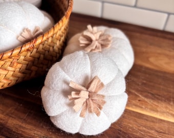 Può includere: Zucche di feltro bianco con steli beige sono esposte su una superficie di legno, alcune in un cestino di vimini. Le zucche hanno un design a coste e sono di un colore neutro. Il cestino è realizzato in materiale intrecciato marrone chiaro.