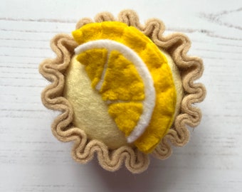 May include: A felt lemon tart with a yellow lemon slice on top. The tart has a brown crust and is made of felt.
