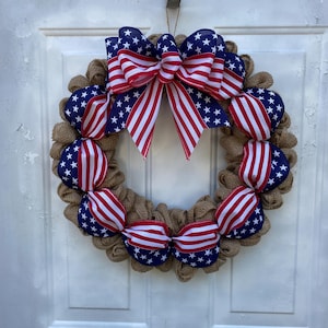 May include: A circular wreath made of natural tan hessian mesh, accented with sections of ribbon featuring a blue background with white stars and red and white stripes. A large, multi-looped bow in the same patriotic ribbon sits at the top centre.