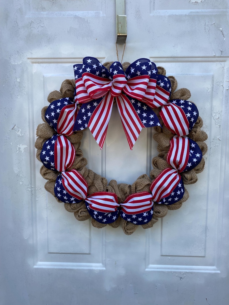 May include: A circular wreath made of natural tan hessian mesh, accented with sections of ribbon featuring a blue background with white stars and red and white stripes. A large, multi-looped bow in the same patriotic ribbon sits at the top centre.