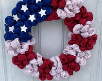 May include: A patriotic wreath with a burlap design. The wreath features blue, red, and white burlap, with white star accents on the blue section. The wreath is designed to hang on a door or wall.