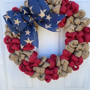 May include: A red, white, and blue burlap wreath with a star-patterned bow. The wreath is made of burlap and is decorated with red and tan burlap ribbons.