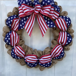 May include: A circular wreath constructed from natural tan hessian mesh, featuring a large decorative bow at the top and smaller ribbon accents throughout. The ribbons display a pattern of red and white stripes alongside a blue field with white stars.
