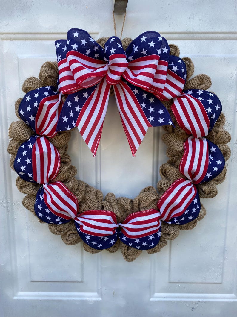 May include: A circular wreath constructed from natural tan hessian mesh, featuring a large decorative bow at the top and smaller ribbon accents throughout. The ribbons display a pattern of red and white stripes alongside a blue field with white stars.