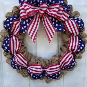 May include: A circular wreath constructed from natural tan burlap mesh, featuring decorative ribbon accents in a patriotic American flag pattern of red, white, and blue stars and stripes, topped with a large, multi-looped bow of the same fabric.