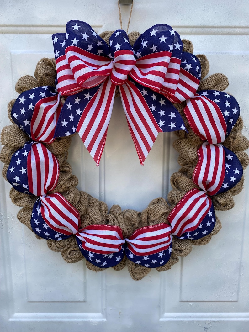 May include: A circular wreath constructed from natural tan burlap mesh, featuring decorative ribbon accents in a patriotic American flag pattern of red, white, and blue stars and stripes, topped with a large, multi-looped bow of the same fabric.