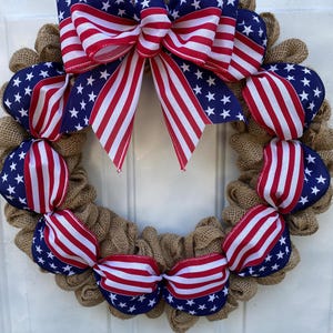 May include: A circular wreath constructed from natural tan burlap mesh, featuring a large decorative bow at the top and smaller ribbon accents throughout. The ribbons display a pattern of white stars on a blue field and red and white stripes.
