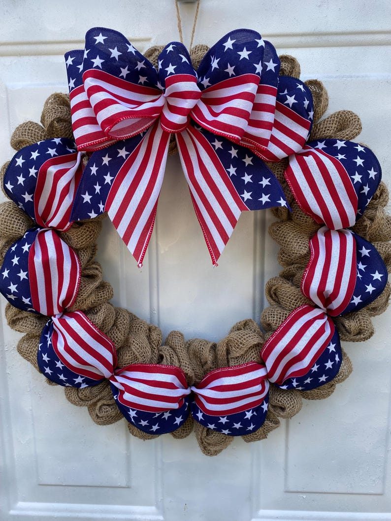 May include: A circular wreath constructed from natural tan burlap mesh, featuring a large decorative bow at the top and smaller ribbon accents throughout. The ribbons display a pattern of white stars on a blue field and red and white stripes.