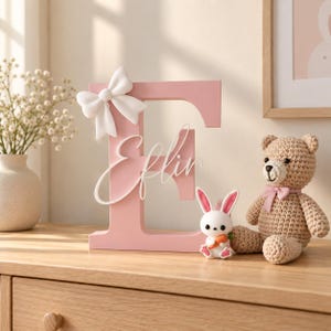 May include: A light pink wooden letter E featuring a white bow attached to the top left corner and the name Eflin written in white cursive script across the center. A small white rabbit figurine holding a carrot sits next to the letter.