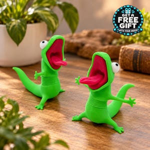 May include: Two bright green lizard figurines with wide-open mouths, protruding pink tongues, and large white eyes. Each lizard features a ribbed midsection and a long tail. A circular badge in the corner displays the text GET A FREE GIFT WITH YOUR ORDER.