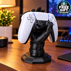 May include: A black robotic hand stand holding a white and black video game controller. The stand features a segmented wrist design on a hexagonal base. A circular badge in the upper right corner displays the text GET A FREE GIFT WITH YOUR ORDER.