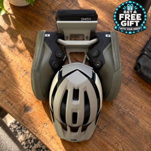 May include: A light grey bicycle helmet positioned in front of a light grey wall-mounted storage rack. Two olive green cycling shoes with black dials are attached to the rack, and a small black Smith branded case sits on the top shelf of the rack.