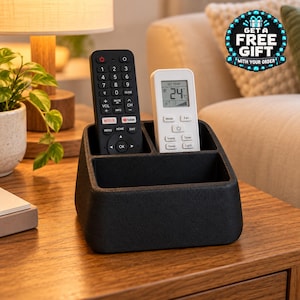 Pode incluir: Um organizador de mesa preto fosco com três compartimentos, contendo um controle remoto de televisão preto e um controle remoto de ar condicionado branco com um display digital mostrando 24 graus Celsius. O organizador tem um acabamento texturizado.