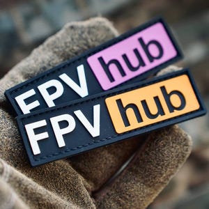 Puede incluir: Dos parches rectangulares con bordes negros y letras blancas. El parche superior presenta el texto FPV hub con un fondo rosa detrás de la palabra hub. El parche inferior presenta el texto FPV hub con un fondo naranja detrás de la palabra hub.