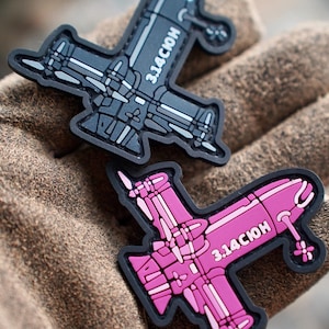 Könnte beinhalten: Zwei gummierte Aufnäher in Form stilisierter Flugzeuge, einer in Dunkelgrau und einer in leuchtendem Pink. Beide Aufnäher haben schwarze Umrandungen und den weißen Schriftzug 3.14СЮН auf dem Rumpf. Die Designs zeigen detaillierte Flügel- und Triebwerksmarkierungen.