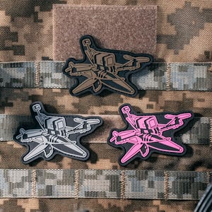 Puede incluir: Tres parches tácticos con diseño de dron, cada uno con un borde negro. Un parche es marrón, otro gris claro y otro rosa brillante. Los parches se muestran sobre una tela de camuflaje junto a un dispositivo negro con la etiqueta VJ UK.