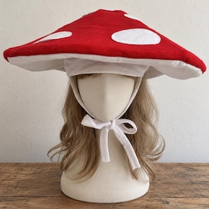 May include: A wide-brimmed hat shaped like a mushroom cap, featuring a vibrant red top with white circular felt spots. The underside of the brim is white, and the hat includes white fabric chin ties that are knotted securely under the chin.